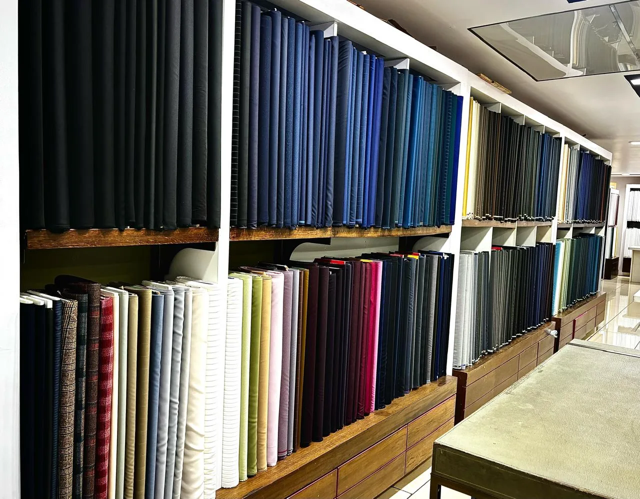 A wall of suiting rolls at Bespoke Studio — navy and black on the top shelf with solids, tweeds and checks below