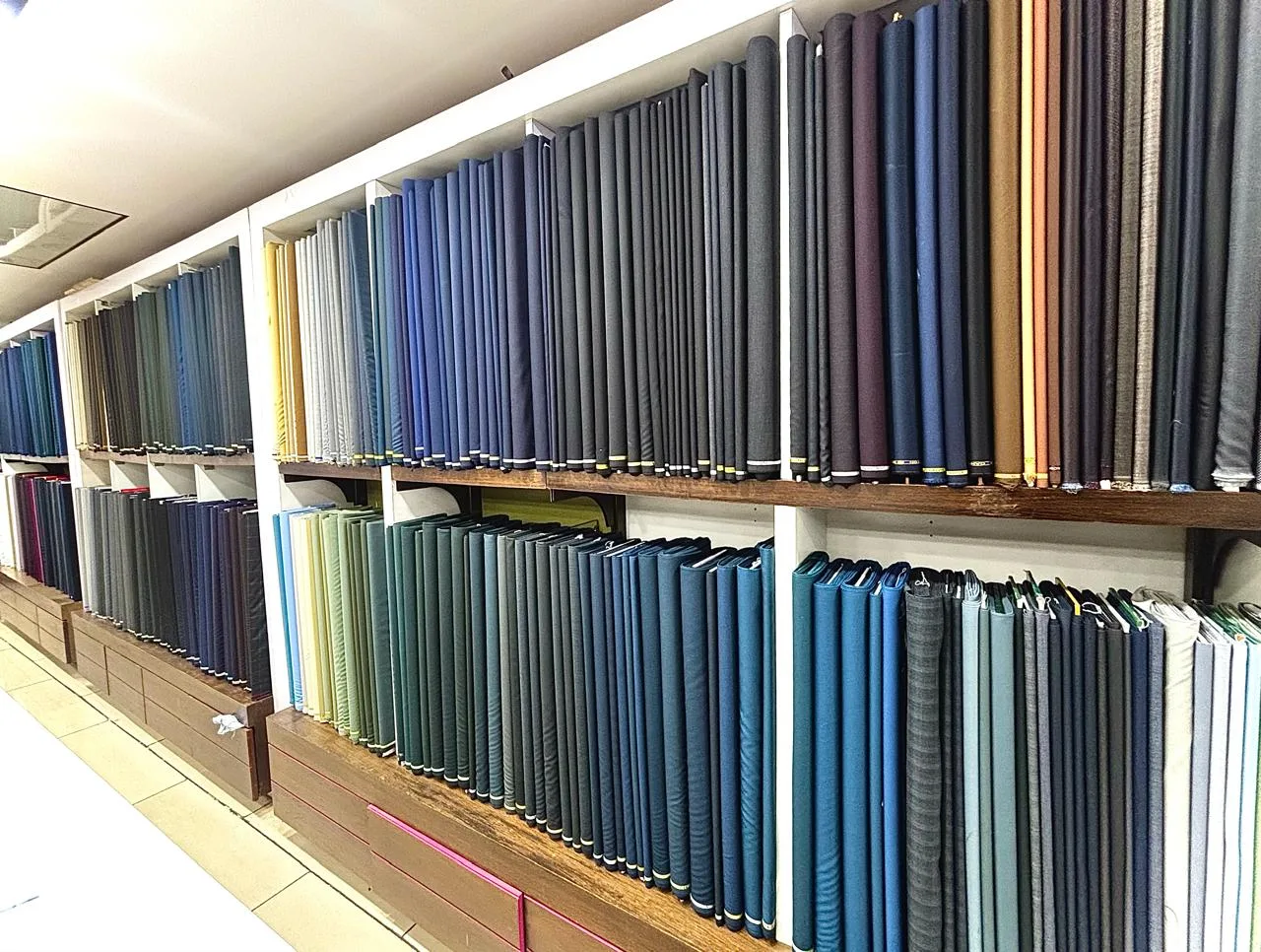 A full wall of suiting fabric rolls in greys, blues and browns, densely stacked at the Bespoke Studio showroom