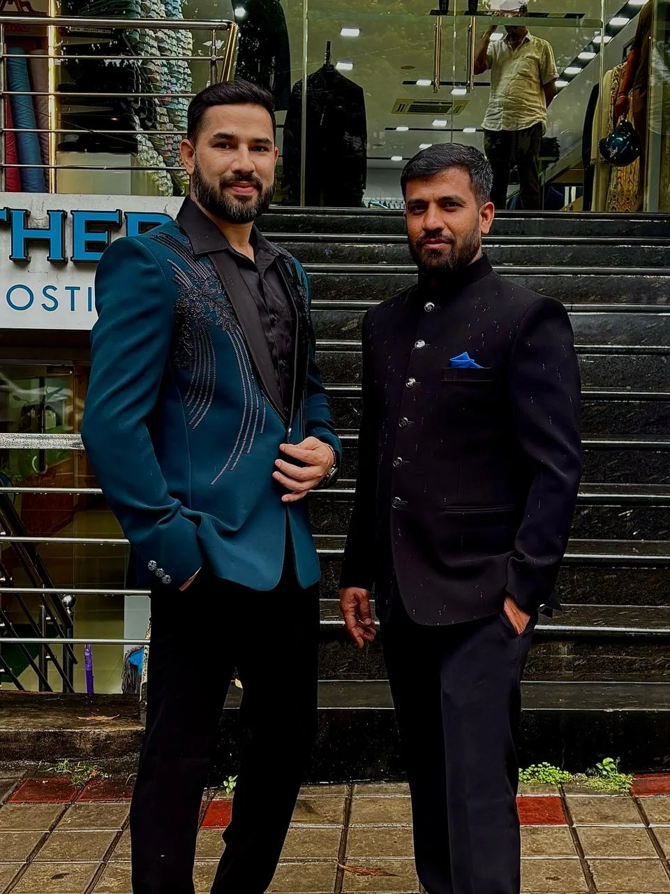 Two customers on the showroom steps — one in a teal embellished tuxedo, one in a black bandhgala with a blue pocket square