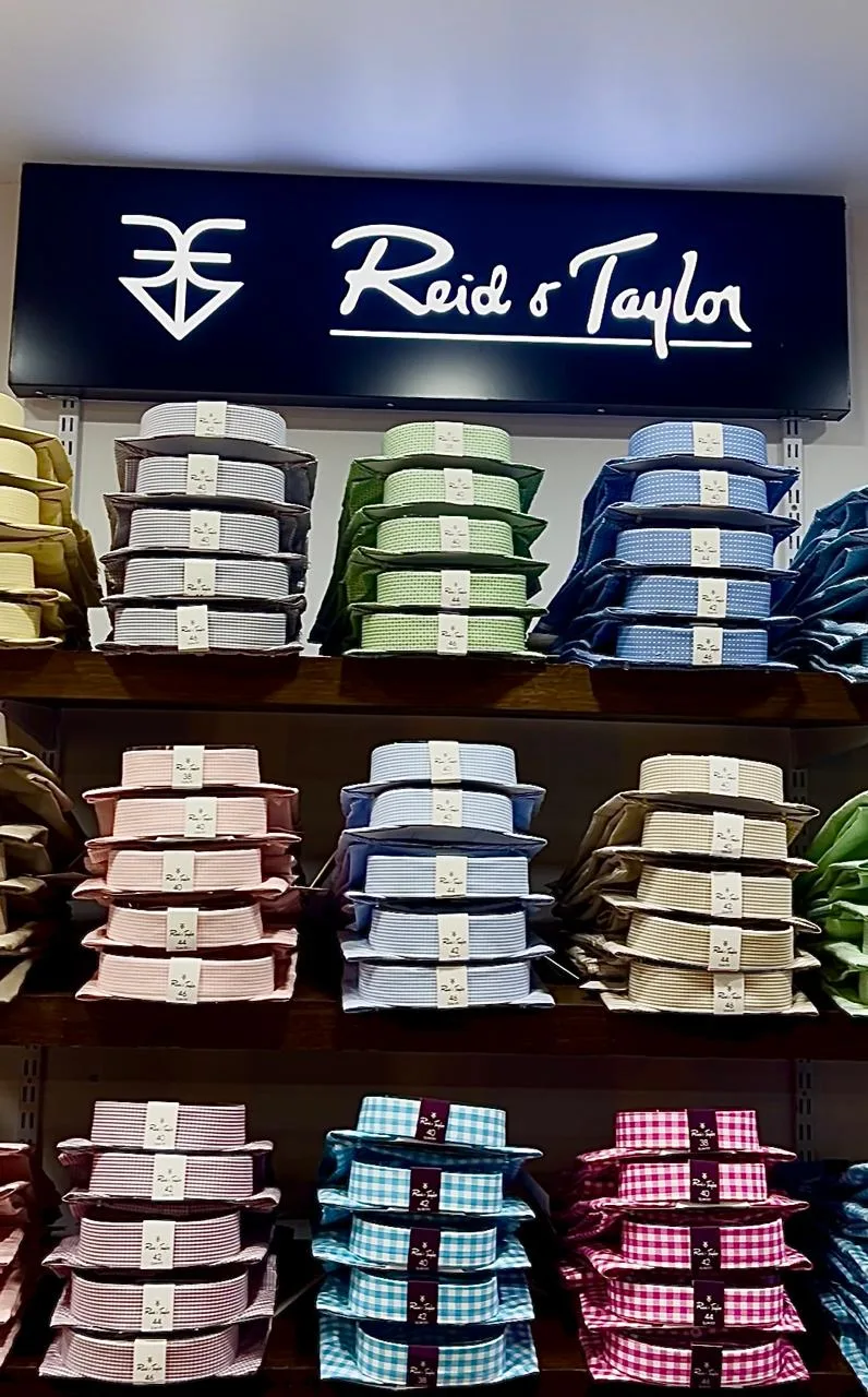 Reid & Taylor shirting display — gingham and check shirt pieces stacked in size-tagged boxes beneath the navy Reid & Taylor signboard