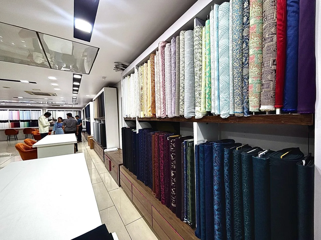 A wide aisle at the Bespoke Studio showroom — colourful ethnic and jacquard fabric rolls on the upper shelves, dark suiting below, with customers browsing near the orange chairs