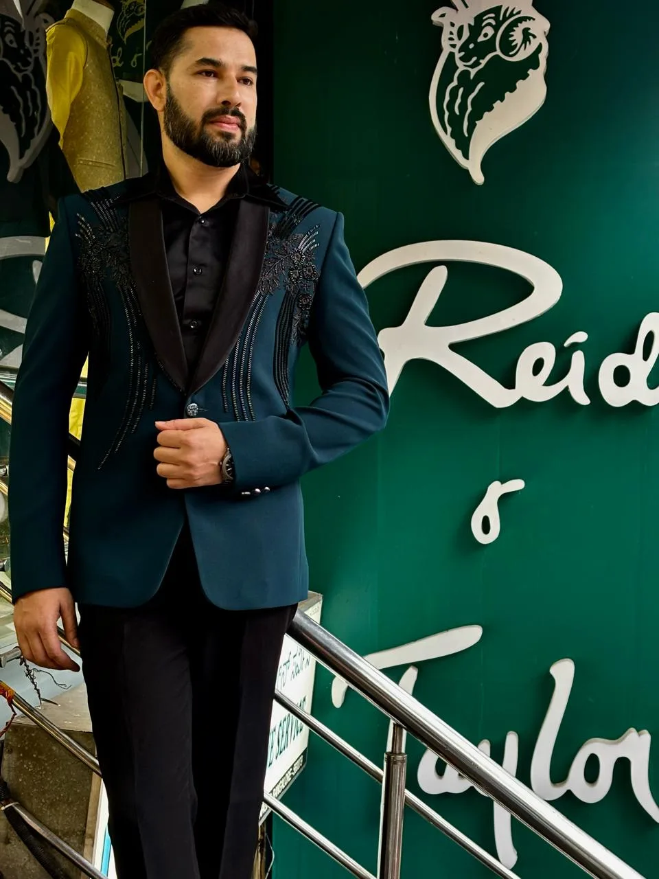 Customer in a teal embellished tuxedo blazer with a black shawl lapel before the green Reid & Taylor signboard